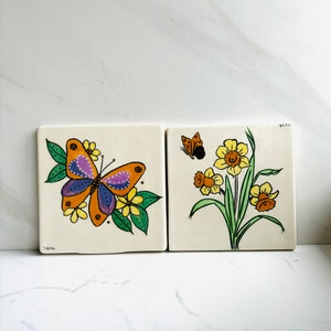 May include: Two square ceramic tiles with hand-painted designs. The first tile features a yellow butterfly with purple and blue accents on a green leafy branch with yellow flowers. The second tile features a cluster of yellow daffodils with orange centers and a small orange butterfly. Both tiles are signed "Vera".
