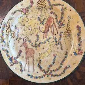 May include: A decorative ceramic plate featuring hand-painted African animals, including giraffes, elephants, and deer. The plate has a cream-colored background with colorful floral and leaf accents. The rim is green.