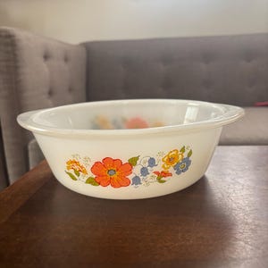 May include: White oval serving bowl with a floral design. The bowl features a pattern of orange, yellow, and blue flowers. The bowl is sitting on a dark brown wooden surface.