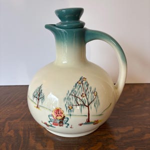 May include: A vintage ceramic pitcher with a teal lid and a hand-painted scene of a couple sitting on a bench under a willow tree. The pitcher has a round body and a handle.