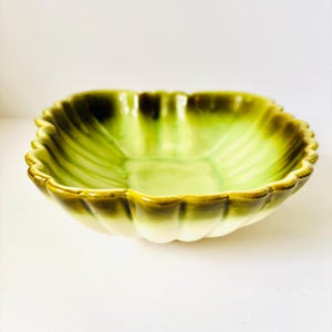 May include: A square ceramic bowl with a scalloped rim. The bowl features a gradient of green hues, transitioning from a light green interior to a darker green and brown rim. The bowl has a ribbed texture.
