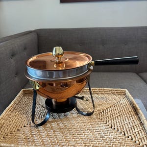 May include: A copper-toned fondue pot with a black handle and lid, set on a black stand. The pot sits on a woven rectangular tray. The lid has a gold-colored knob. The pot is designed for cooking and serving.
