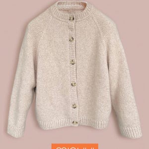 Puede incluir: Cárdigan beige claro con cuello redondo y cierre de botones. El suéter de punto tiene mangas largas. El nombre de la marca "minuui" se muestra en un rectángulo naranja en la parte inferior. El cárdigan está hecho de un material de punto suave.