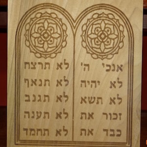 May include: Wooden plaque with engraved Hebrew text and decorative circular designs. The text is arranged in two columns and includes the Ten Commandments. The wood has a light, natural finish.