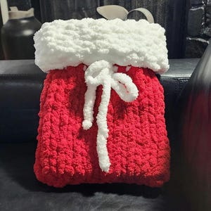 Könnte beinhalten: Ein handgefertigter Weihnachtsstrumpf in Rot mit weißem, flauschigem Bund. Ein weißer Krawattenverschluss ist mittig auf der Vorderseite angebracht. Der Strumpf ist aus dickem, strukturiertem Garn gefertigt.