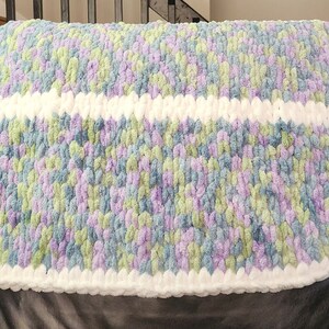 Puede incluir: Una manta suave de crochet con un borde blanco y un patrón multicolor de azul, verde y morado.