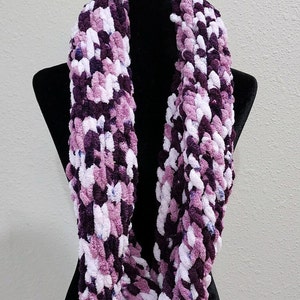May include: A purple, white, and pink infinity scarf made of fuzzy yarn. The scarf is displayed on a black mannequin.
