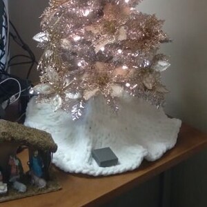 May include: A white faux fur tree skirt with a gray box underneath. The tree skirt is decorated with a gold and white Christmas tree.