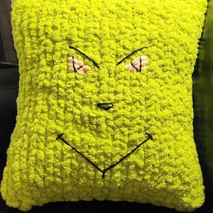 May include: A lime green, square-shaped decorative pillow with a stitched face. The face features black stitched eyebrows, a nose, and a smile. The eyes have a peach-colored accent.