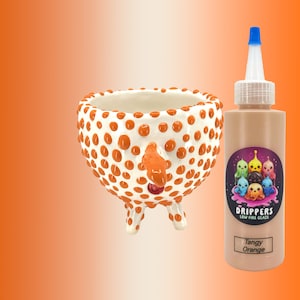 May include: A white ceramic bowl with orange polka dots and three white legs. Next to it is a bottle of "Drippers" low fire glaze in "Tangy Orange" color, with a blue nozzle and a colorful label.