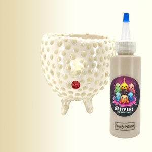 May include: A bottle of Pearly White low fire glaze for ceramics. The bottle is white with a colourful cartoon label featuring a group of dripping characters. The glaze is made by Drippers.
