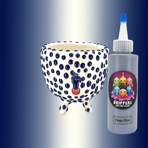 Puede incluir: Una maceta de cerámica blanca con lunares azules y un pico rojo. La maceta tiene tres patas y está decorada con un esmalte azul. Una botella de esmalte de baja temperatura Deep Blue está al lado de la maceta. La botella tiene un diseño de dibujos animados de personajes coloridos.