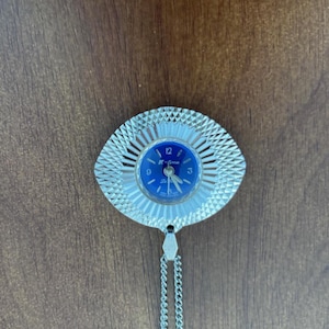 May include: A vintage silver-toned pendant watch with a blue face. The watch has a circular face with the numbers 12, 3, and 9 visible. The watch is attached to a silver chain. The watch face has the text "K-time".
