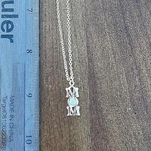May include: A silver-colored necklace with a pendant spelling "MOM". The pendant has a small, round, light-colored stone in the center of the "O". The necklace is on a wooden surface.
