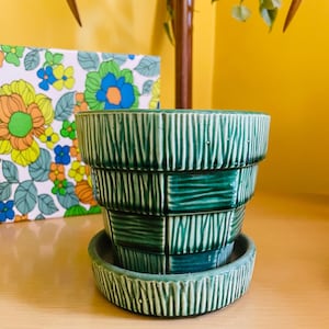 Könnte beinhalten: Ein grüner Vintage-Keramik-Blumentopf mit passender Untertasse. Der Topf hat ein mehrschichtiges Design mit vertikalen und horizontalen strukturierten Linien. Ein farbenfroher Blumendruck ist im Hintergrund zu sehen. Der Topf ist für den Innenbereich.