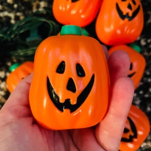 May include: A string of orange plastic pumpkin-shaped light covers, each featuring a carved jack-o'-lantern face with black painted eyes, a triangular nose, and a wide grinning mouth. The pumpkins are attached to a dark green electrical cord.