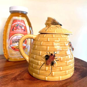 May include: A ceramic honey pot shaped like a beehive, with a lid and handle. The pot is yellow with a textured design and painted bees. A bottle of honey with a label that reads "Raw Honey" is in the background.