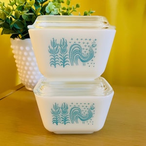 Op de afbeelding: Twee gestapelde vierkante voorraadbakjes van wit melkglas met doorzichtige glazen deksels. Elk bakje heeft een blauwgroene grafische print van een haan naast maïskolven, geaccentueerd door kleine kruisvormige symbolen boven de haan.