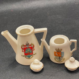 May include: Two miniature ceramic teapots with lids. The teapot on the left has a gold trim and a crest with a tree and the text "Mayor of Colwyn". The teapot on the right has a gold trim and a crest with a crown and the text "Ramsgate".