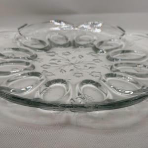 May include: Clear glass egg serving platter with a scalloped edge. The platter has a central section for dip and twelve egg indentations. The glass has a floral design etched into the center.