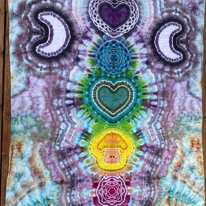 May include: A tie-dye textile with a vibrant, psychedelic design. The artwork features symmetrical patterns including crescent shapes, hearts, and a central mushroom motif. The color palette includes shades of purple, blue, green, and yellow, creating a visually striking piece.
