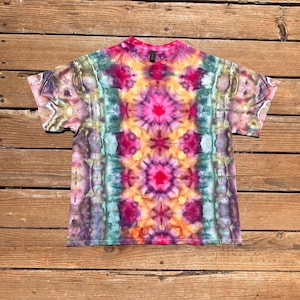 May include: A short-sleeve crew neck t-shirt featuring a vibrant tie-dye pattern with a symmetrical design. The color palette includes bright pink, deep magenta, golden yellow, teal, and muted purple tones arranged in a repeating mandala-like motif.