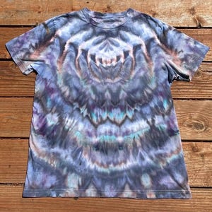 May include: A short-sleeved t-shirt with a tie-dye pattern in shades of blue, purple, and teal. The design features concentric circles radiating from the center, creating a unique and artistic look. The shirt is made of soft fabric.