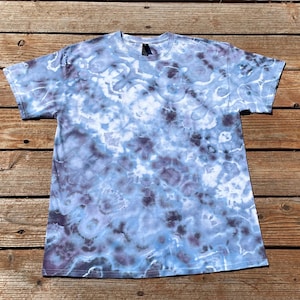 May include: A short-sleeve crew neck t-shirt featuring a tie-dye pattern in shades of light blue, dark blue, and white. The fabric displays a marbled, cloud-like effect with blended dark and light tones distributed across the entire garment surface.