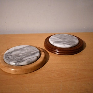 May include: Two round marble coasters with wooden bases. The coasters are grey and white with a marbled pattern. The bases are made of light brown wood.