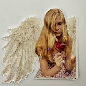 Puede incluir: Una pegatina que presenta a una persona con cabello largo y rubio, sosteniendo una manzana roja. La persona lleva un vestido floral y tiene alas de ángel. Las alas y el cabello tienen detalles de purpurina en forma de estrella.