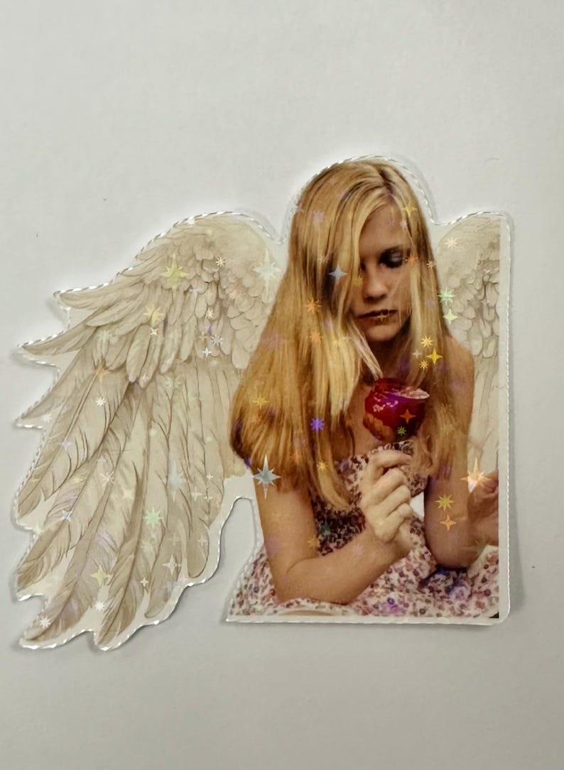 May include: A sticker featuring a person with long blonde hair, holding a red apple. The person is wearing a floral dress and has angel wings. The wings and hair have star-shaped glitter accents.