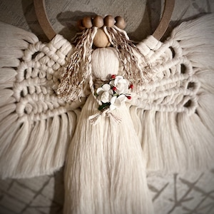 May include: A white macrame angel with a wooden head and a floral crown. The angel has long, flowing hair and a white dress with a floral embellishment.