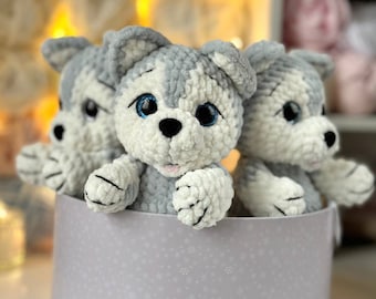 May include: Three gray and white crocheted husky dog plushies with blue eyes are sitting in a round, light purple box with a snowflake pattern.