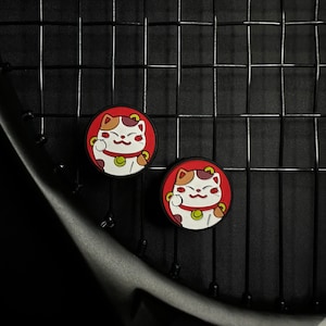 Puede incluir: Dos amuletos de gato de la suerte de color rojo, blanco y marrón con ojos negros en relieve y una moneda de oro sobre un fondo de raqueta de tenis negra.