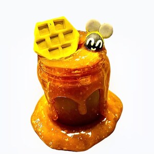 May include: A glass jar filled with honey, dripping down the sides. A yellow waffle and a cartoon bee are on top of the jar.