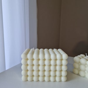 Könnte beinhalten: Zwei weiße Kerzen in Säulenform mit einem strukturierten, blasenartigen Design. Die Kerzen stehen auf einer weißen Oberfläche.