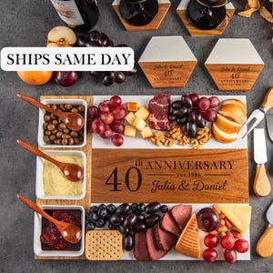 May include: A rectangular wooden and white serving board engraved with 40th ANNIVERSARY est. 1986 Julia & Daniel, featuring three square white bowls with wooden spoons, two hexagonal coasters, two wine glasses, a bottle, and cheese knives.
