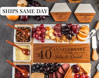 May include: A rectangular wooden and white serving board engraved with 40th ANNIVERSARY est. 1986 Julia & Daniel, featuring three square white bowls with wooden spoons, two hexagonal coasters, two wine glasses, a bottle, and cheese knives.