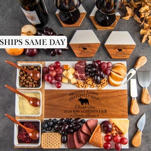 May include: A rectangular wooden and white serving board with the text PROUDLY RAISED GRATEFULLY SOLD 2026 IOWA COUNTY FAIR Brody Lorenz, three white square bowls with wooden spoons, grapes, cheese, crackers, meat, nuts, and cheese knives.