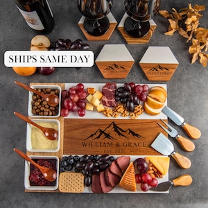 May include: A charcuterie board with a wooden and marble design, featuring the text "WILLIAM & GRACE EST. 2021". The board is filled with various cheeses, meats, grapes, nuts, and crackers. Includes wooden serving spoons and cheese knives. Also includes wine glasses and coasters.