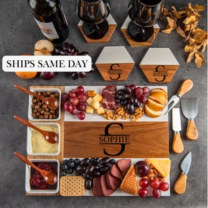 May include: A wooden charcuterie board with white marble and wood sections, filled with grapes, cheese, and meats. Includes small white bowls with wooden spoons, cheese knives, and personalized coasters. The board is engraved with the name "SOPHIE".
