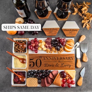May include: A charcuterie board with a white and wood grain design, filled with various cheeses, meats, grapes, nuts, and crackers. The board is engraved with "50th Anniversary" and the names "Doris & Larry". Includes wine glasses, coasters, and serving utensils.