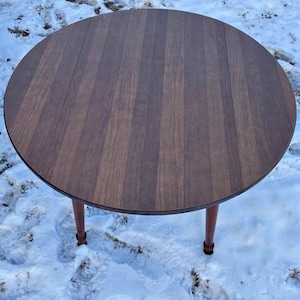 May include: A round wooden table with a brown wood grain finish. The table has a leaf that can be extended to make it larger. The table is sitting on a snowy surface.