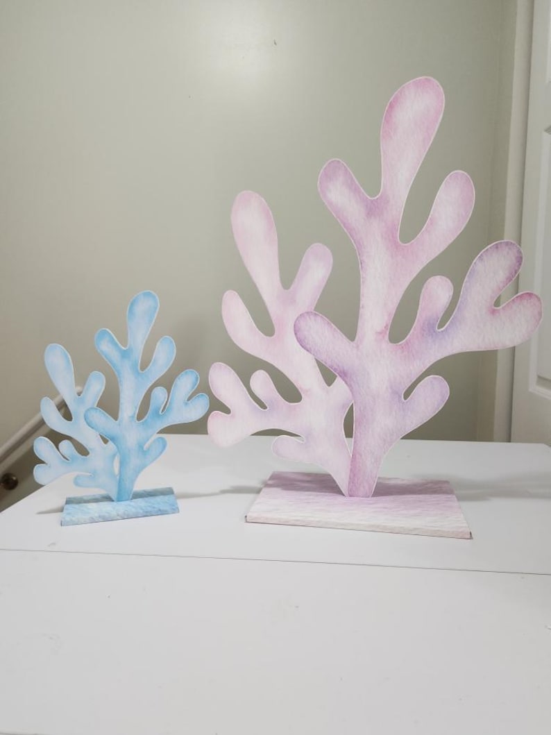 May include: Two decorative paper sea coral pieces, one blue and one pink, on a white surface. The coral is made of paper and has a watercolor effect.