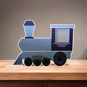 May include: A flat, train-shaped picture frame in shades of light and dark blue. The locomotive design features a square photo opening in the cabin, a tall smokestack, and four black wheels along the bottom edge, all outlined in a white border.