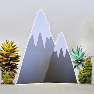 May include: A gray paper mountain wall decor with a white outline. The mountain is made of two peaks, one larger than the other. The mountain is surrounded by small green plants.