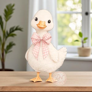 May include: A charming, cream-colored duck figurine with a pink gingham bow around its neck. The duck has orange feet and a matching beak. The figurine is standing on a wooden surface, with a blurred background of a window and greenery.