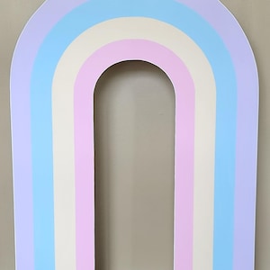 May include: A large, freestanding arch shaped like a rainbow, featuring vertical stripes in pastel shades of lavender, light blue, pale yellow, and soft pink. The structure has a smooth, flat surface and a clean, arched top design.
