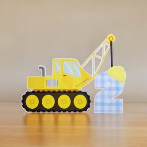 May include: A yellow construction crane toy lifting a blue and white checkered number two with a yellow hard hat on top. The crane has black tracks and a yellow cab.