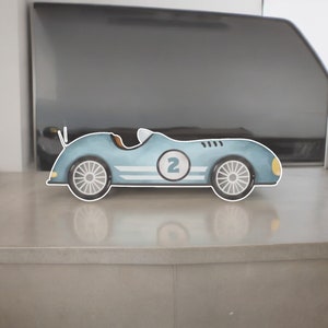 May include: A light blue toy race car with a white number 2 inside a circle on the side. The car features white racing stripes, a small white windshield, yellow headlights, and grey spoked wheels. It has a sleek, low-profile design.